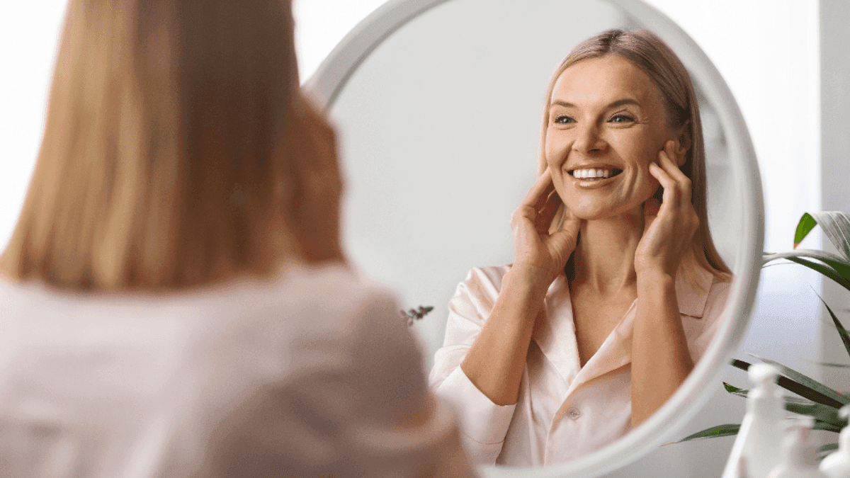 Woman smiling at her reflection in a round mirror while gently touching her face